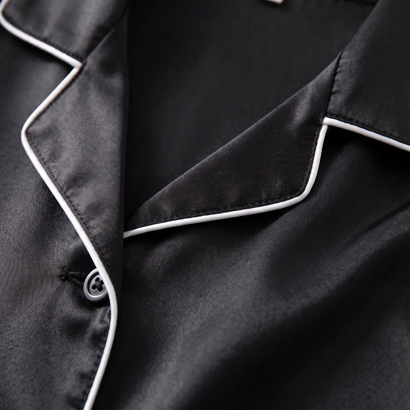 Close-up of the notched collar of the black satin pyjama shirt, with white piping, stitching and a grey button. 5
