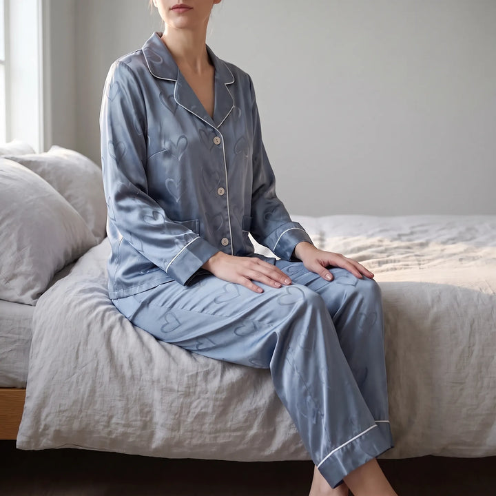 Woman sitting on the edge of a bed in blue satin pyjamas with a tonal heart pattern and white piping. 1