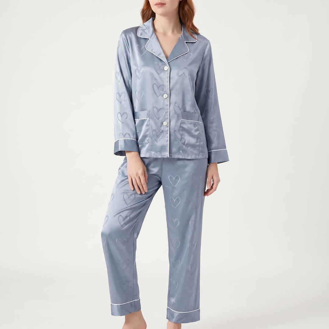 Model standing in blue satin pyjamas with heart jacquard pattern, front view showing buttons, two chest pockets and white piping. 2