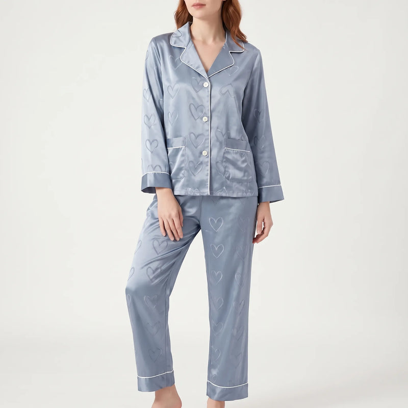 Model standing in blue satin pyjamas with heart jacquard pattern, front view showing buttons, two chest pockets and white piping. 2
