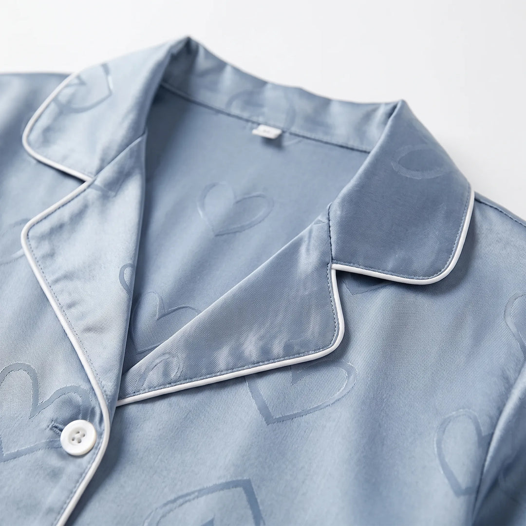 Close-up of the blue satin pyjama top's notched collar with white piping, a white button and the tonal heart pattern. 5