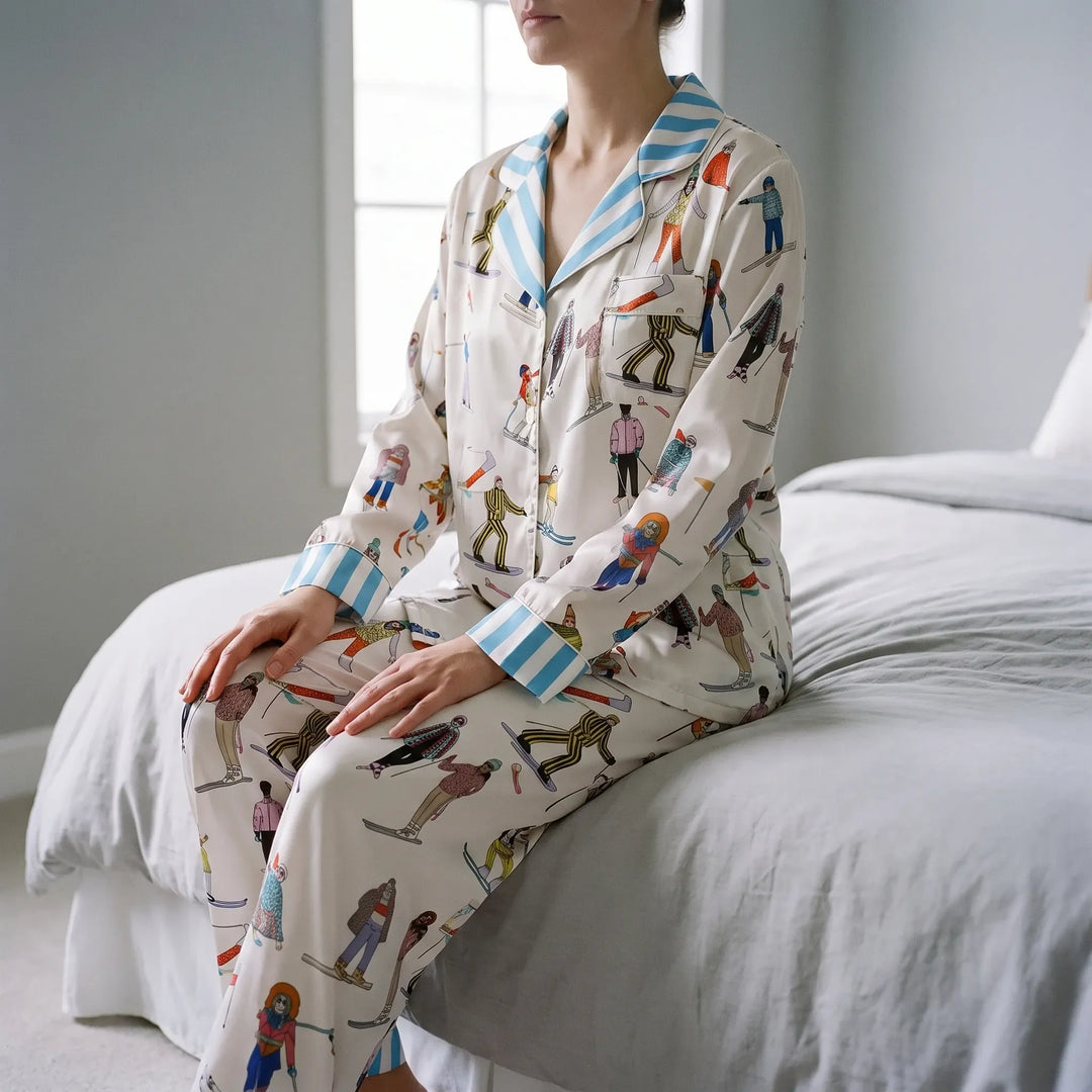 Model sitting on the edge of a bed in a cream satin pyjama set with skier illustrations and blue striped collar and cuffs. 1