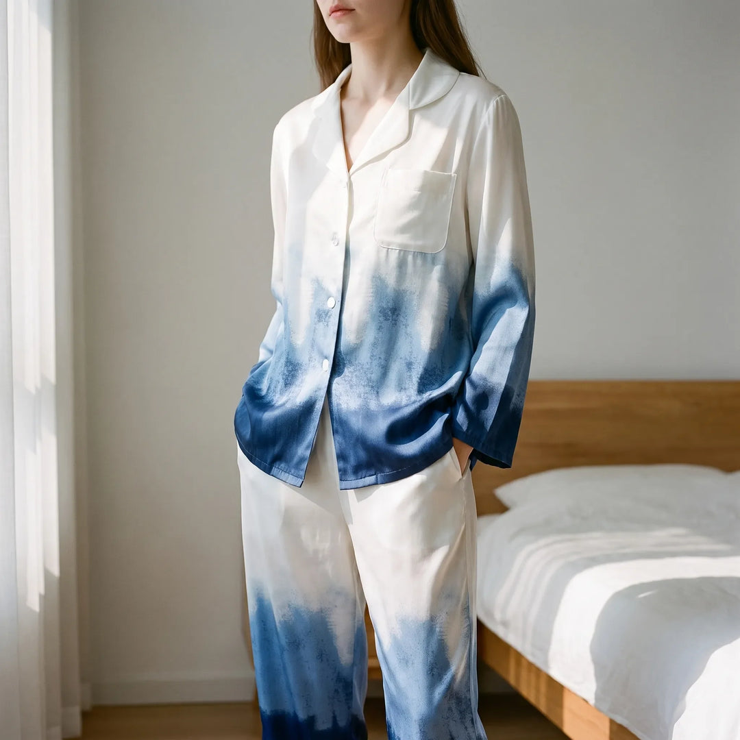 Model in white-to-blue ombre satin pyjamas standing in a sunlit bedroom beside a wooden bed, hands in pockets. 1