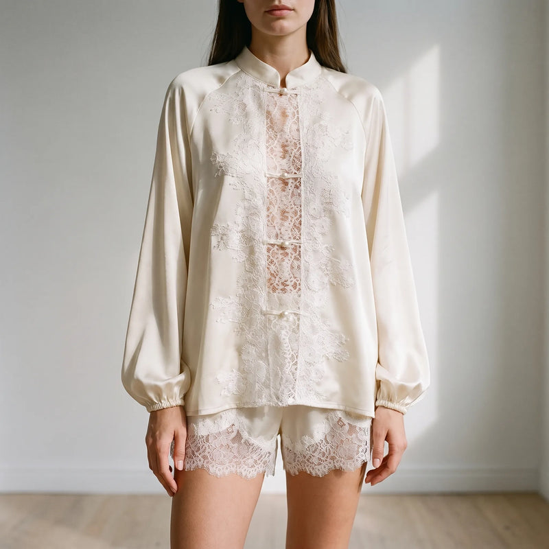 Model in a cream satin pyjama set with a lace panel down the front, pearl buttons and lace-trimmed shorts, in a bright room. 1