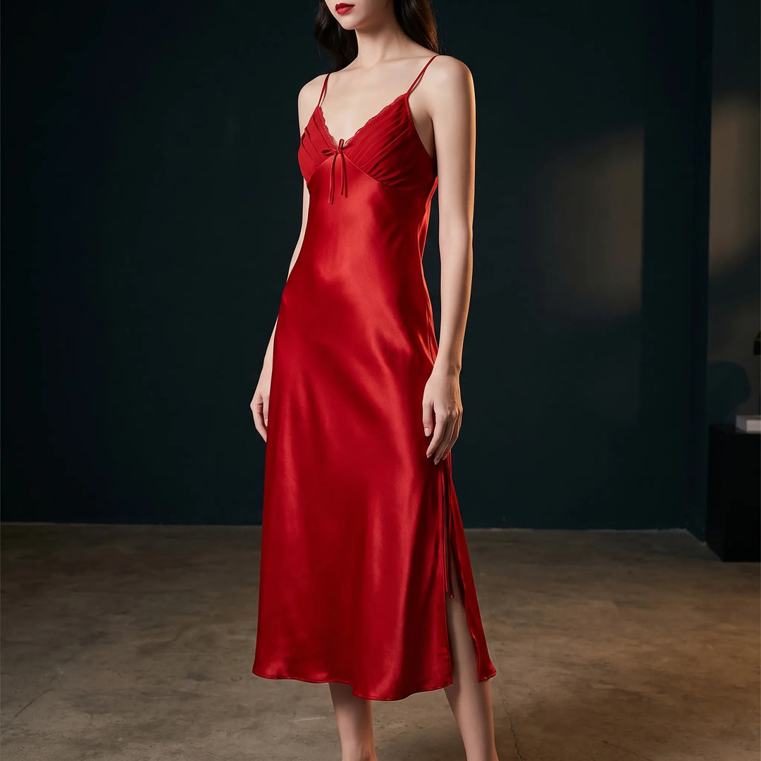 Model in a red satin slip nightdress with pleated lace-trimmed bodice, bow and side slit, posed against a dark studio backdrop. 1