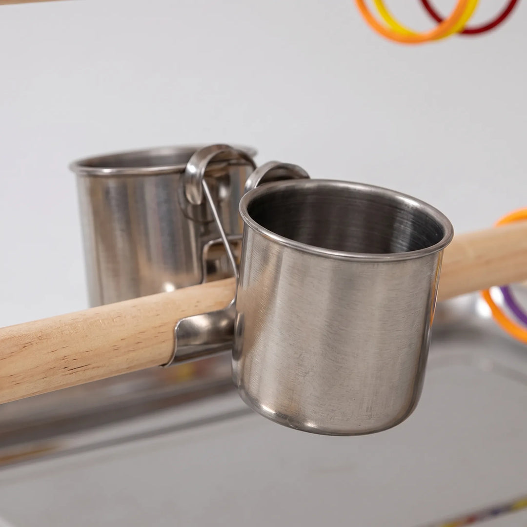 Close-up of two stainless steel feeding cups clipped onto a wooden perch above the metal tray, with coloured rings nearby. 3