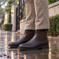 Brown Chelsea-style rain boots covered in raindrops, worn with beige trousers on a wet pavement beside a garden wall. 1