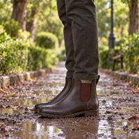 Muddy brown Chelsea rain boots worn with olive trousers on a puddled leafy park path, with water droplets on the surface. 3
