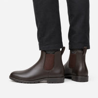 Side view of brown Chelsea rain boots with elastic gussets, worn with dark grey trousers against a plain white background. 4