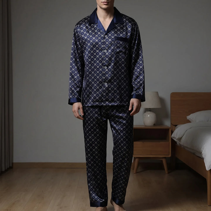 Model wearing the navy satin pyjama set with a diamond lattice print, standing barefoot in a bedroom beside a bed and lamp. 1