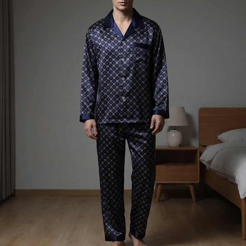 Model wearing the navy satin pyjama set with a diamond lattice print, standing barefoot in a bedroom beside a bed and lamp. 1