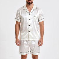 Front view of white satin pyjama shirt and shorts with black piping, worn by a bearded model against a plain light background. 2