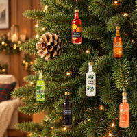 Six colorful miniature glass bottle ornaments with gold caps hanging on a lit evergreen branch beside a pine cone in a cozy room. 1