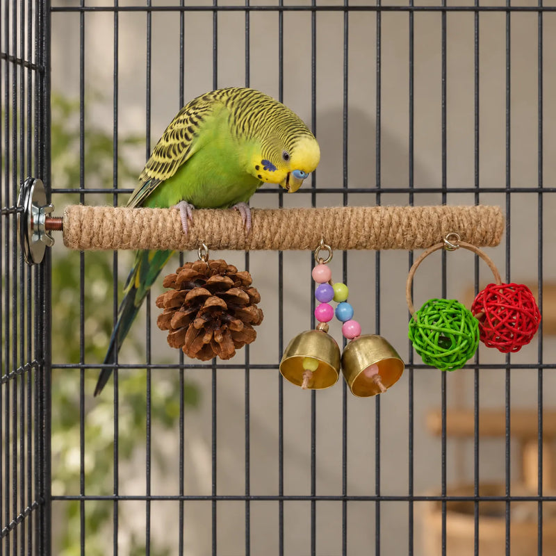 Green and yellow budgerigar perched on the rope-wrapped bar inside a cage, looking down at the hanging pine cone, bells and woven balls. 2