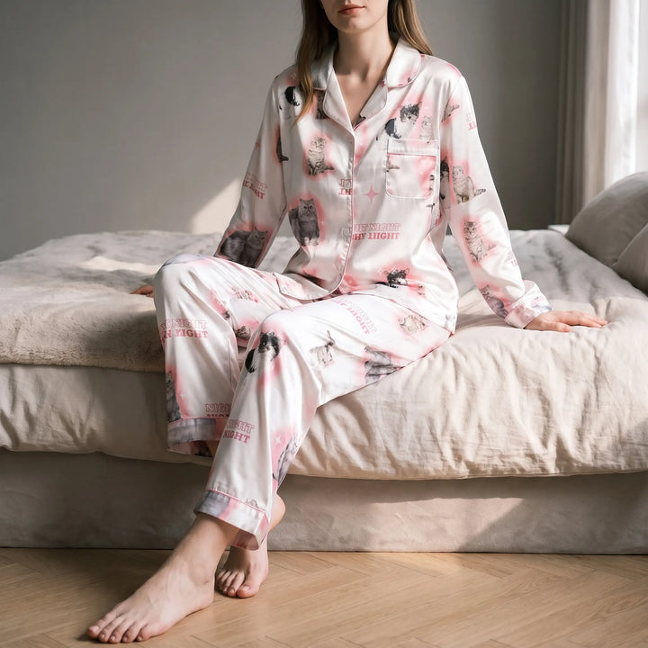Woman sitting barefoot on the edge of a bed in the white satin pyjama set with pink-glow kitten print. 1