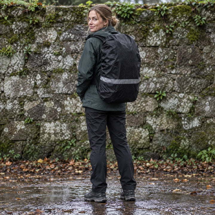 Woman looking over her shoulder wearing a black rain cover with a reflective band on her rucksack, standing on a wet path by a stone wall. 1