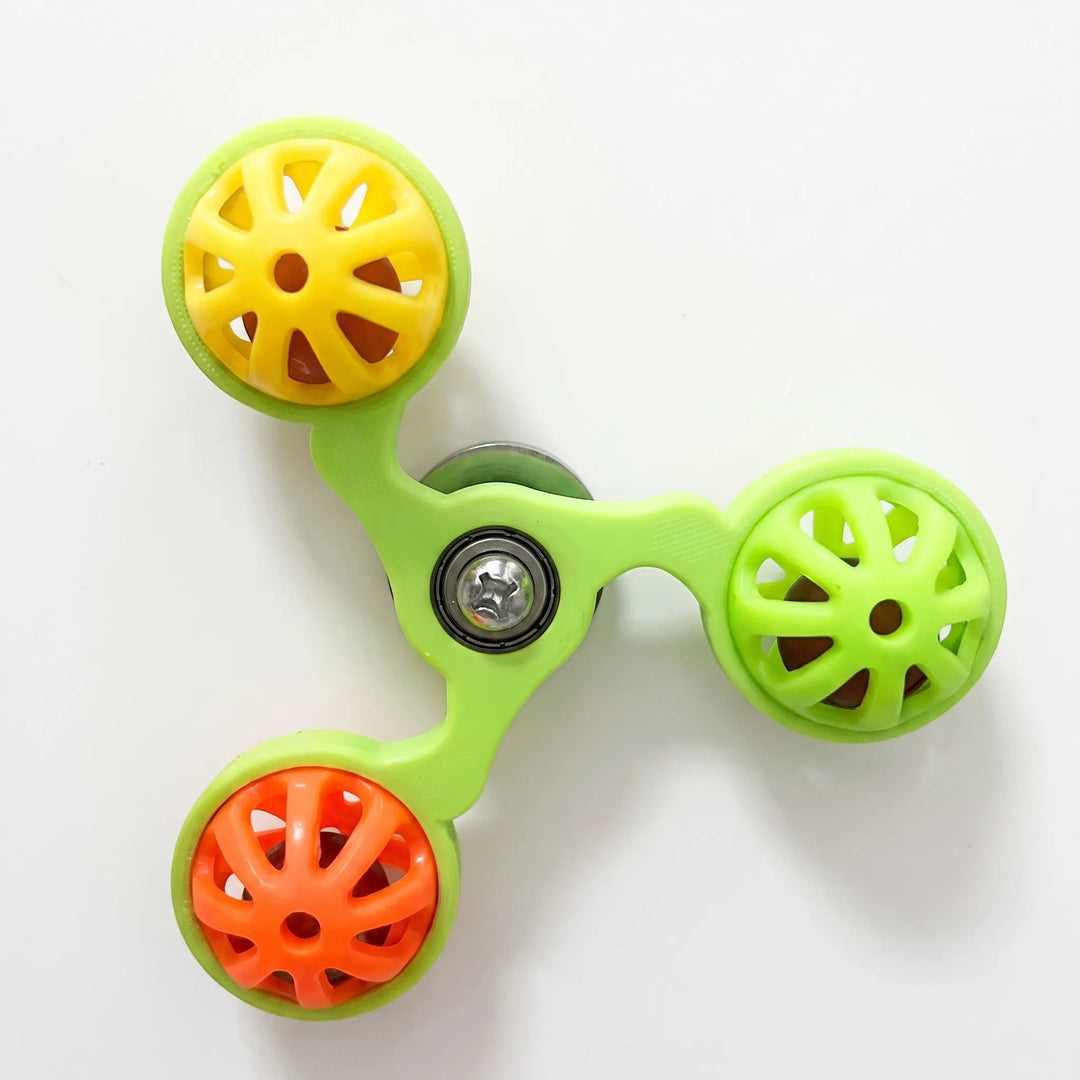 Green three-armed spinner seen from above on a plain white surface, with yellow, lime and orange perforated bell balls around a central bolt. 1