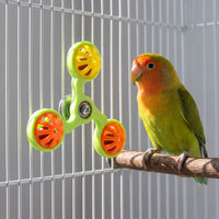 Green spinner with yellow and orange bell balls fixed to a white cage, while a lovebird perches on a wooden branch beside it. 3