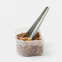 Narrow metal scoop standing in a clear plastic cup filled with mixed seeds and grains, on a plain white background. 1