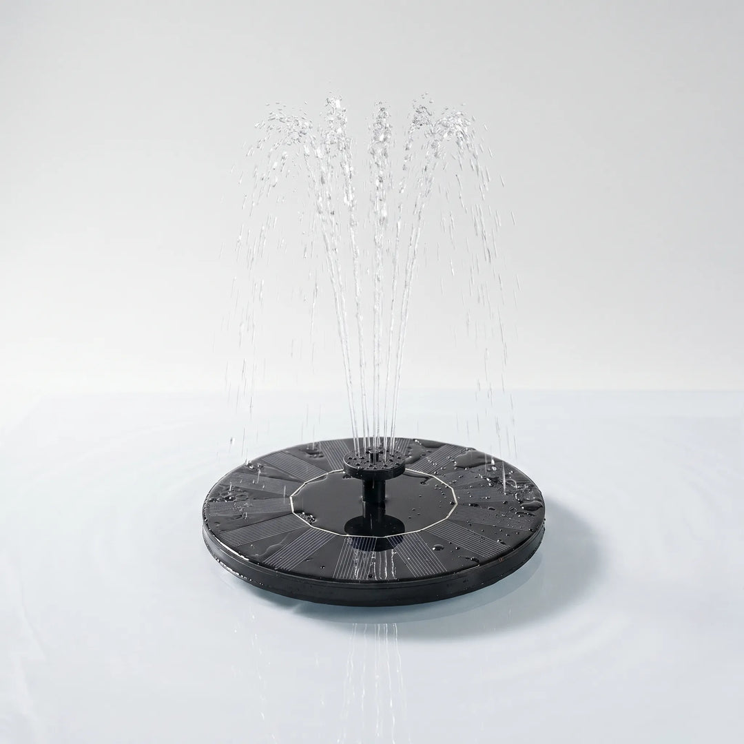 Round black floating solar fountain on still water, spraying a fan of fine jets from its central nozzle against a plain pale background. 1