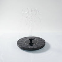 Round black floating solar fountain on still water, spraying a fan of fine jets from its central nozzle against a plain pale background. 1