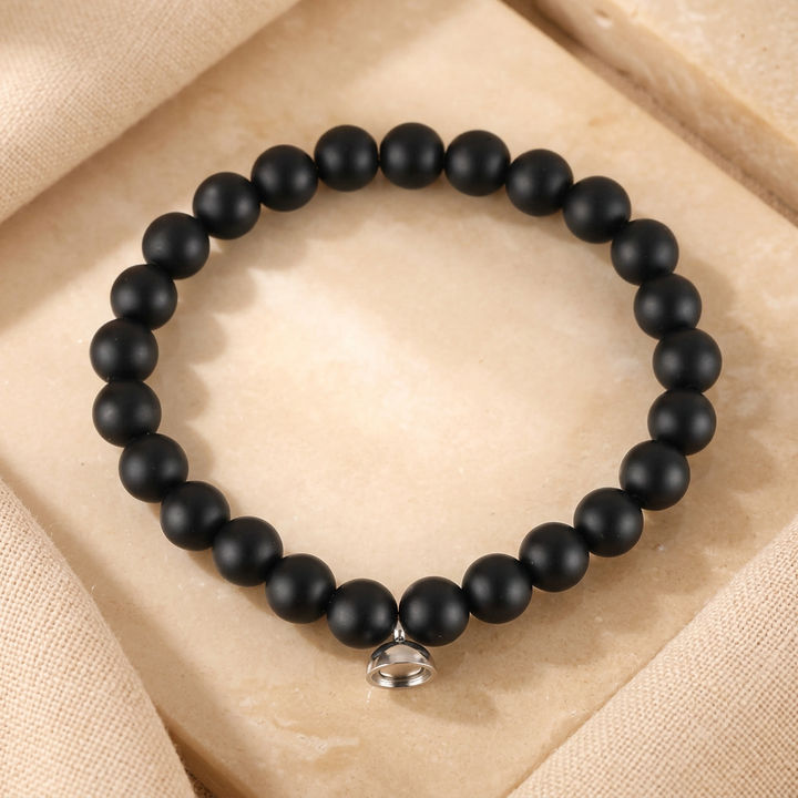 Matte black bead bracelet with a small silver magnetic charm, laid flat on a pale stone tile 2