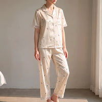 Woman standing in a sunlit room wearing the cream satin-look pyjama set with bow and dot print and lace trim. 1