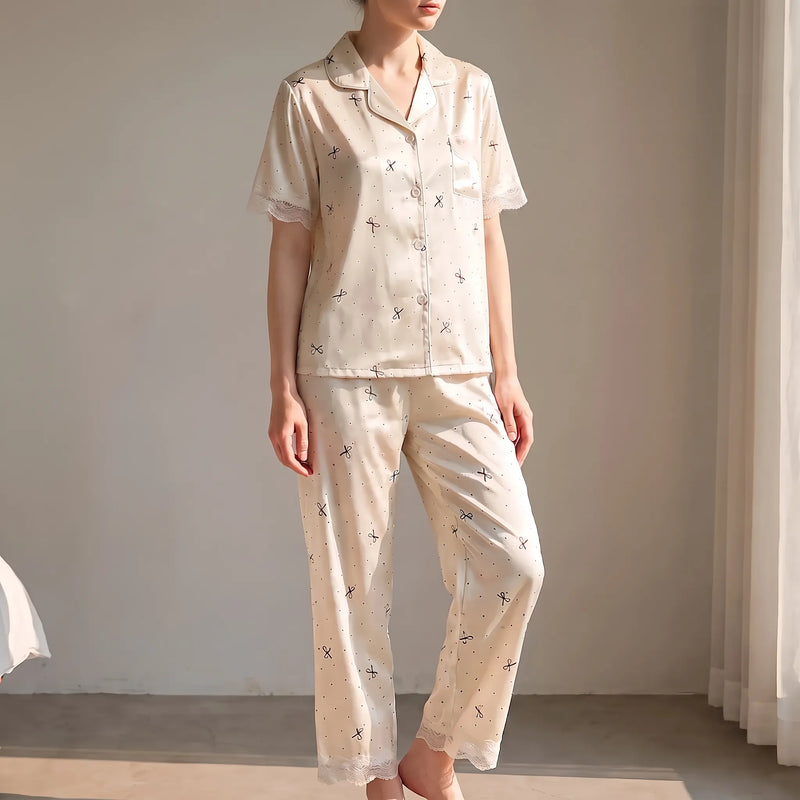 Woman standing in a sunlit room wearing the cream satin-look pyjama set with bow and dot print and lace trim. 1