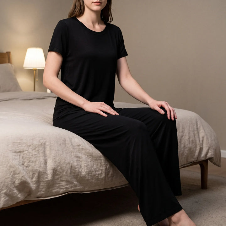 Woman in a black short-sleeved top and wide-leg trousers sitting on the edge of a bed in a softly lit bedroom. 1