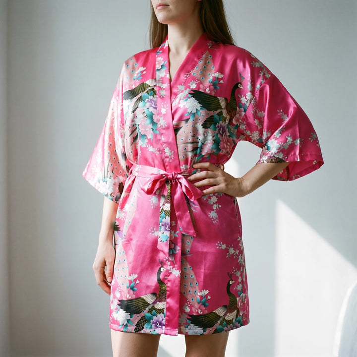 Woman in a satin kimono robe in hot pink with peacock and blossom print, belted at the waist, hand on hip. 1