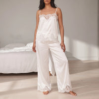 Model in a white satin lace-trimmed camisole and wide-leg trousers standing barefoot in a bright bedroom beside a bed with white linen. 1