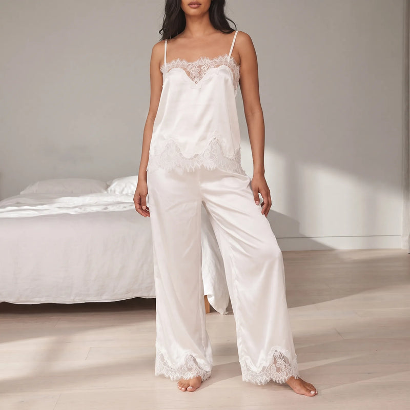 Model in a white satin lace-trimmed camisole and wide-leg trousers standing barefoot in a bright bedroom beside a bed with white linen. 1