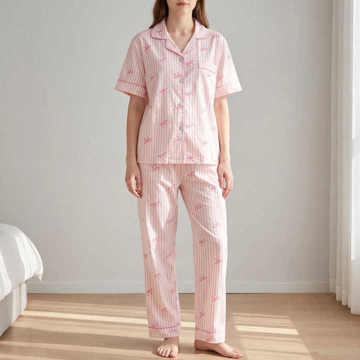 Model standing in a bright bedroom wearing the pink striped bow-print pyjama shirt with chest pocket and matching long trousers. 1