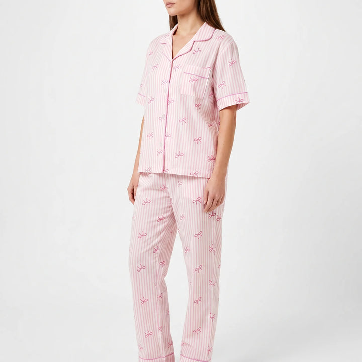 Three-quarter side view of a model wearing the pink striped pyjama shirt with chest pocket and long bow-print trousers. 4
