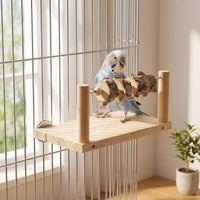 Blue and white budgie standing on the wooden platform inside a wire cage, beside the beaded bark-slice bar, with a plant nearby. 2
