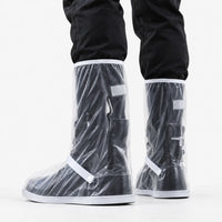 Rear three-quarter view of clear shoe covers with white trim, strap and snap button, worn over black boots against white background. 4