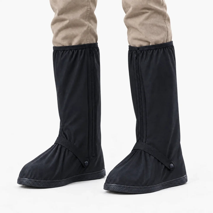 Front view of black tall shoe covers with front zippers and ankle straps worn over khaki trousers on a white background. 7