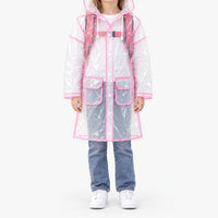 Front view of a girl in the fully transparent raincoat with pink trim, white snap buttons and flap pockets, wearing a pink backpack underneath. 8