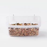 Front view of a clear plastic cage feeder filled with mixed seed, with two metal hooks on top and divided feeding slots. 1