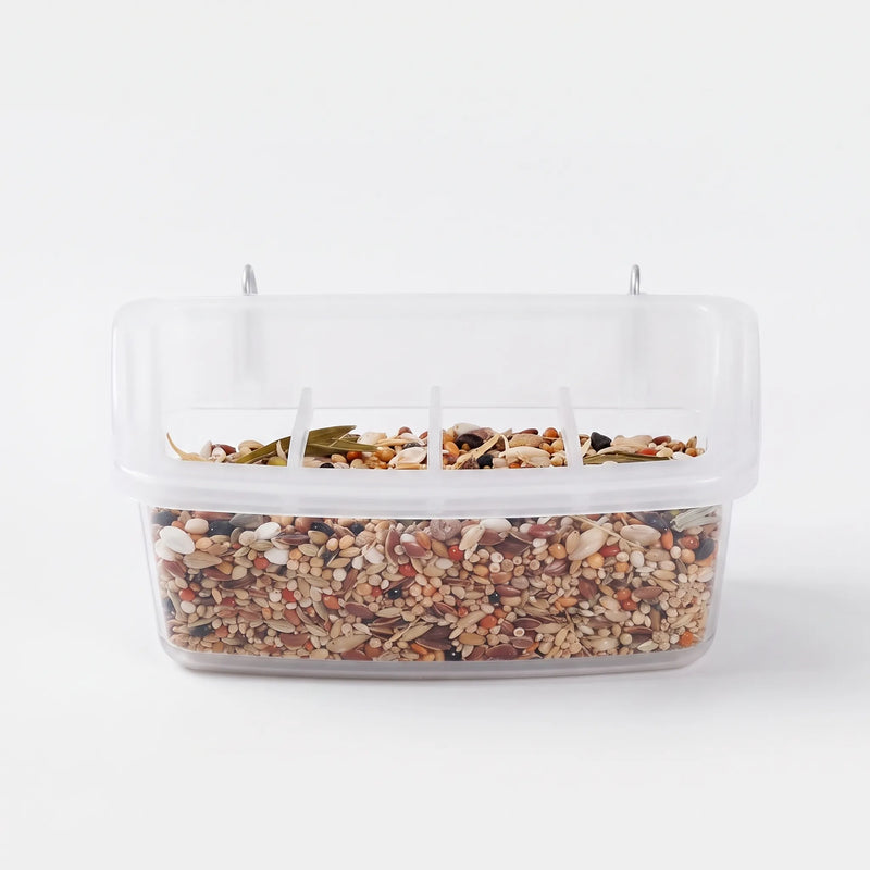 Front view of a clear plastic cage feeder filled with mixed seed, with two metal hooks on top and divided feeding slots. 1