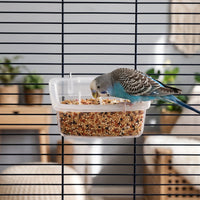 Blue and grey budgerigar perched on the rim of the clear feeder, eating seed, with the feeder hung on a cage. 2