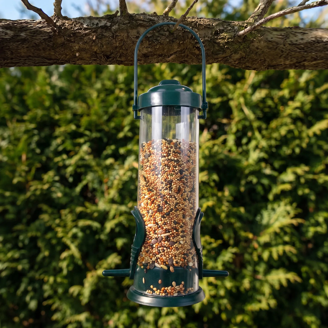 Feeder filled with mixed seed hanging from a tree branch by its green handle, with leafy hedge behind. 2