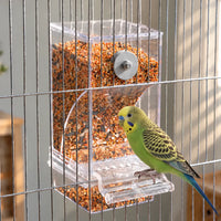 Clear feeder filled with mixed seed, fixed to the bars of a cage with a green budgerigar perched on its tray. 2