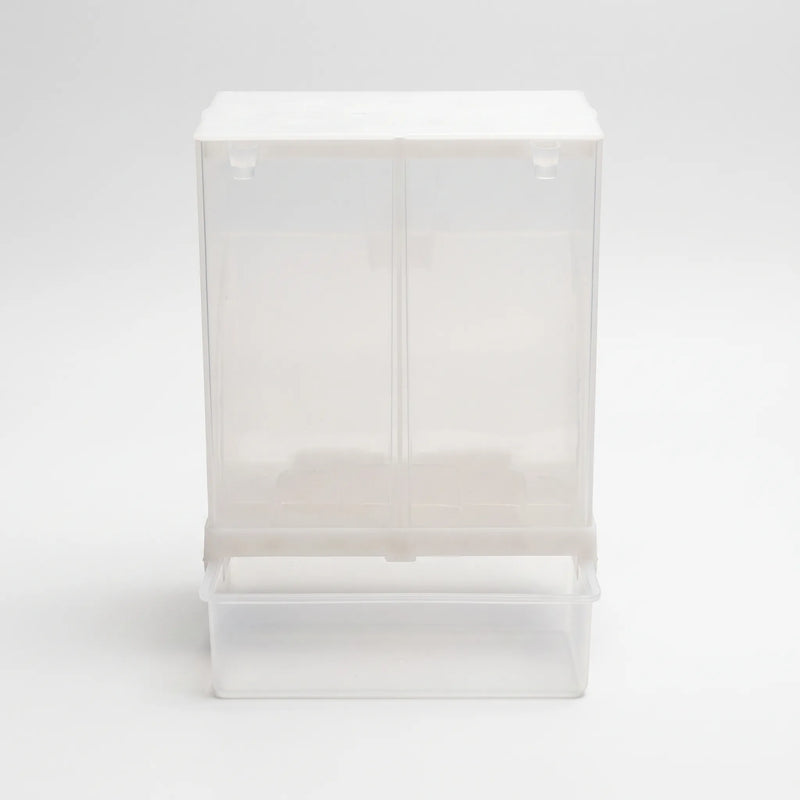 Front view of the clear plastic seed dispenser with its white lid and pull-out tray beneath, shown empty on a plain background. 1
