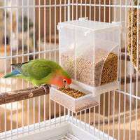 Dispenser hanging on a white cage, filled with seed, while a green lovebird with an orange face perches and eats from the tray. 2
