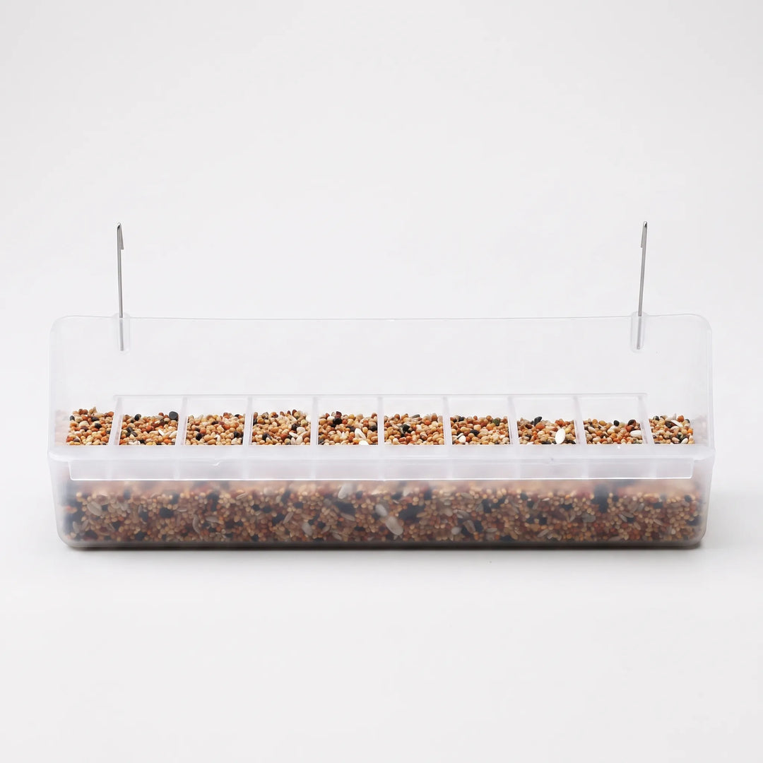 Long clear plastic cage feeder with a row of small seed compartments, filled with mixed seed and two metal hooks on top. 1