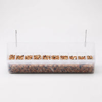 Long clear plastic cage feeder with a row of small seed compartments, filled with mixed seed and two metal hooks on top. 1
