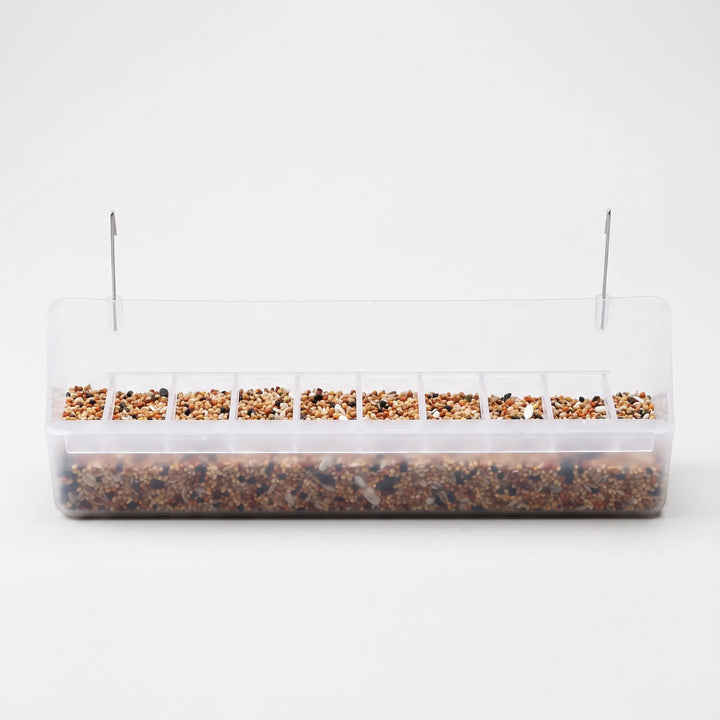 Long clear plastic cage feeder with a row of small seed compartments, filled with mixed seed and two metal hooks on top. 1