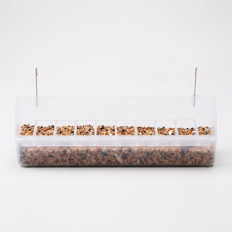 Long clear plastic cage feeder with a row of small seed compartments, filled with mixed seed and two metal hooks on top. 1
