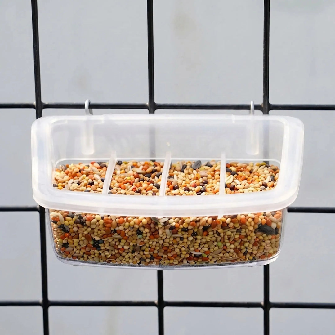 Four-section clear feeder full of mixed seed, hung by two metal hooks on black cage wire. 3